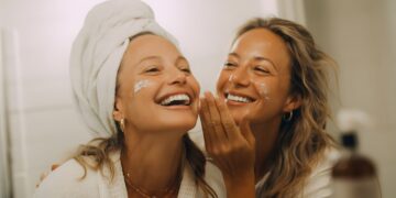 Two young European women applying cold weather skincare moisturizer during winter morning routine with natural window lighting