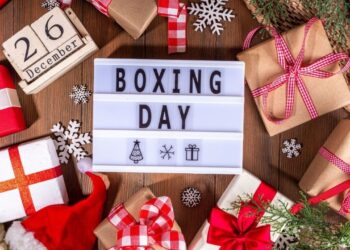 Boxing Day flat lay with wrapped Christmas gifts, festive decorations, snowflakes, and lightbox sign reading Boxing Day on wooden background.