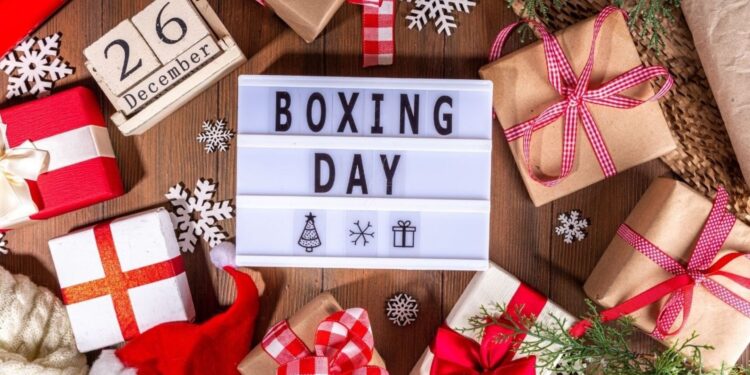 Boxing Day flat lay with wrapped Christmas gifts, festive decorations, snowflakes, and lightbox sign reading Boxing Day on wooden background.