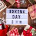 Boxing Day flat lay with wrapped Christmas gifts, festive decorations, snowflakes, and lightbox sign reading Boxing Day on wooden background.