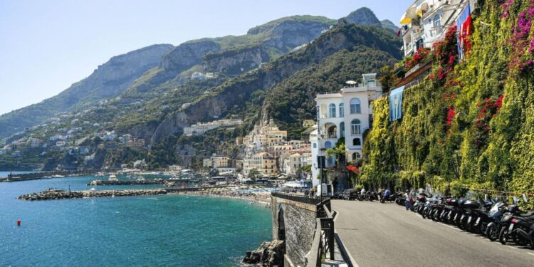 Coastal cliffside town with white buildings, greenery, and a row of scooters along a waterfront road by the blue sea at sea level.