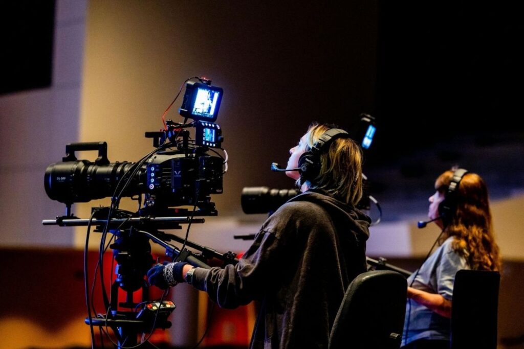 Video camera on a rig with a screen mounted, operator wearing a headset in a dim studio setting