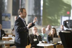 Male speaker in a dark suit gesturing while presenting to a seated audience in a conference room.
