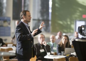 Male speaker in a dark suit gesturing while presenting to a seated audience in a conference room.