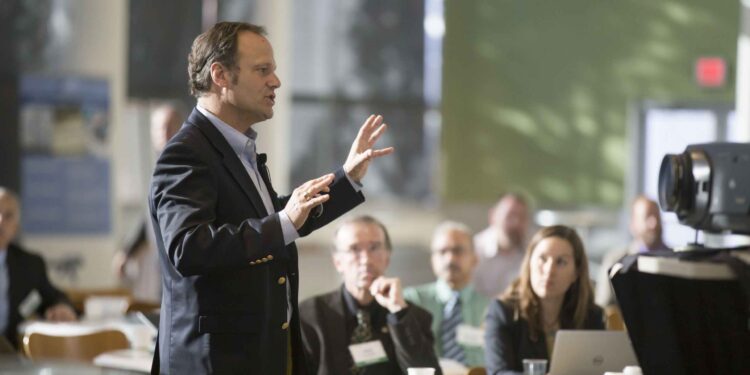 Male speaker in a dark suit gesturing while presenting to a seated audience in a conference room.