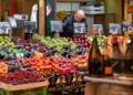 A woman selecting fresh seasonal vegetables at an EU summer farmers market in a sunlit town square.