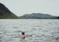 A swimmer wading into a certified EU open-water lake on a clear summer morning, with safety buoys visible in the background.
