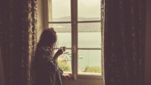 A woman holding a warm cup by a European hotel window, following her morning routine while travelling across EU time zones.