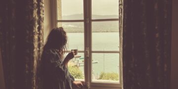 A woman holding a warm cup by a European hotel window, following her morning routine while travelling across EU time zones.