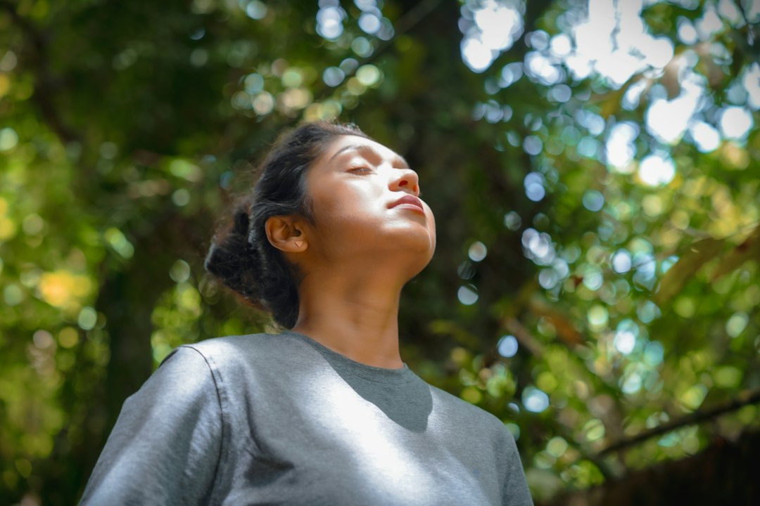 A person stands among tall trees with eyes closed and arms open, fully immersed in a forest bathing experience.
