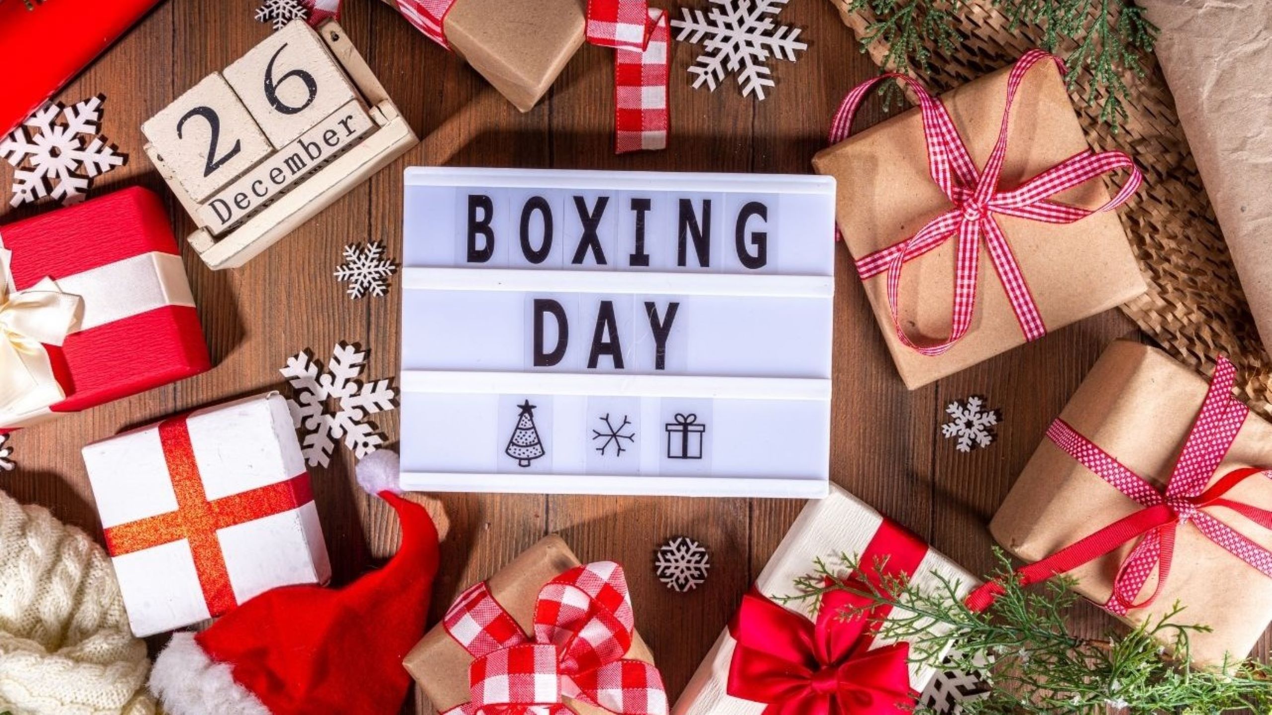 Flat lay do Boxing Day com presentes de Natal embrulhados, decorações festivas, flocos de neve e letreiro luminoso a dizer Boxing Day sobre fundo de madeira.