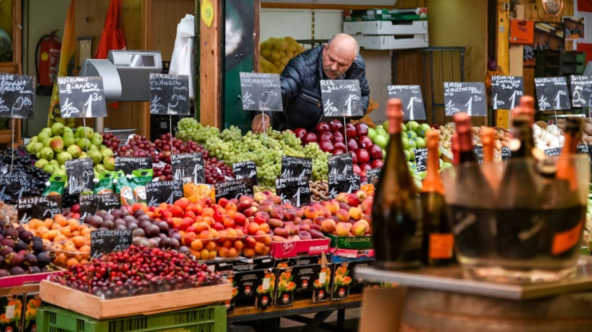 Uma mulher a selecionar vegetais sazonais frescos num mercado de agricultores de verão da UE numa praça de cidade iluminada pelo sol.