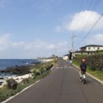 A cyclist on a certified EU coastal cycling route rides along a cliff-top path with the sea visible below