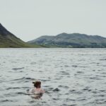 A swimmer wading into a certified EU open-water lake on a clear summer morning, with safety buoys visible in the background.