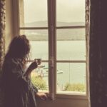 A woman holding a warm cup by a European hotel window, following her morning routine while travelling across EU time zones.